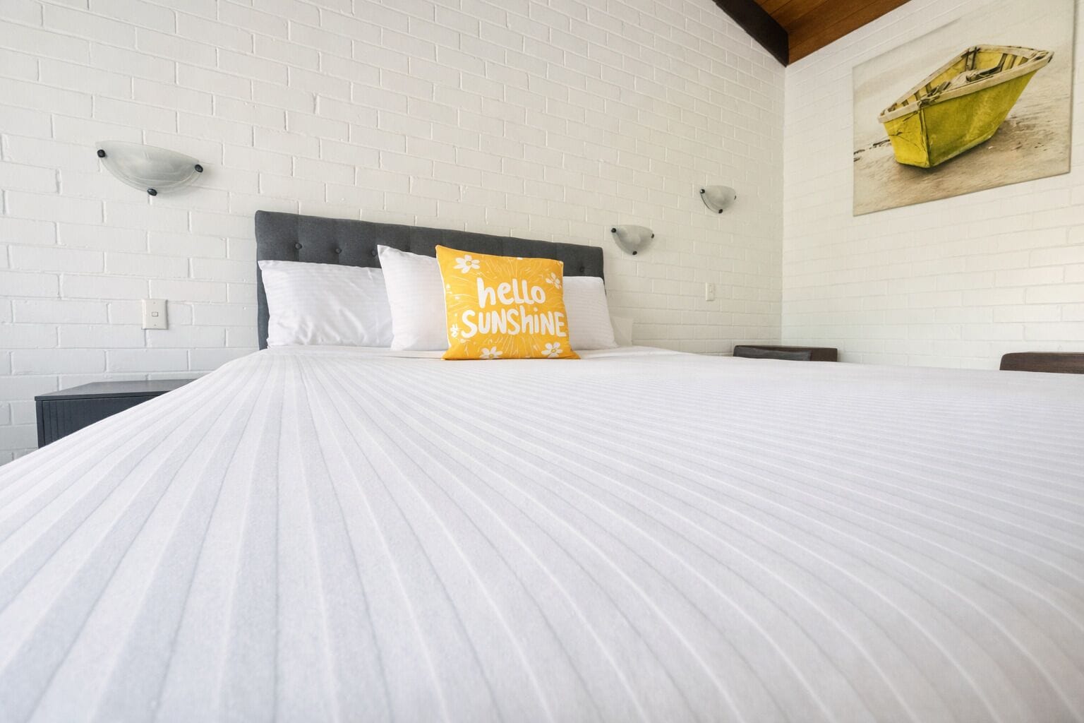 Een hotelkamer met een witte bakstenen muur, een bed met witte beddengoed, een kussen met de tekst 'hello sunshine' en een schilderij van een boot aan de muur.
