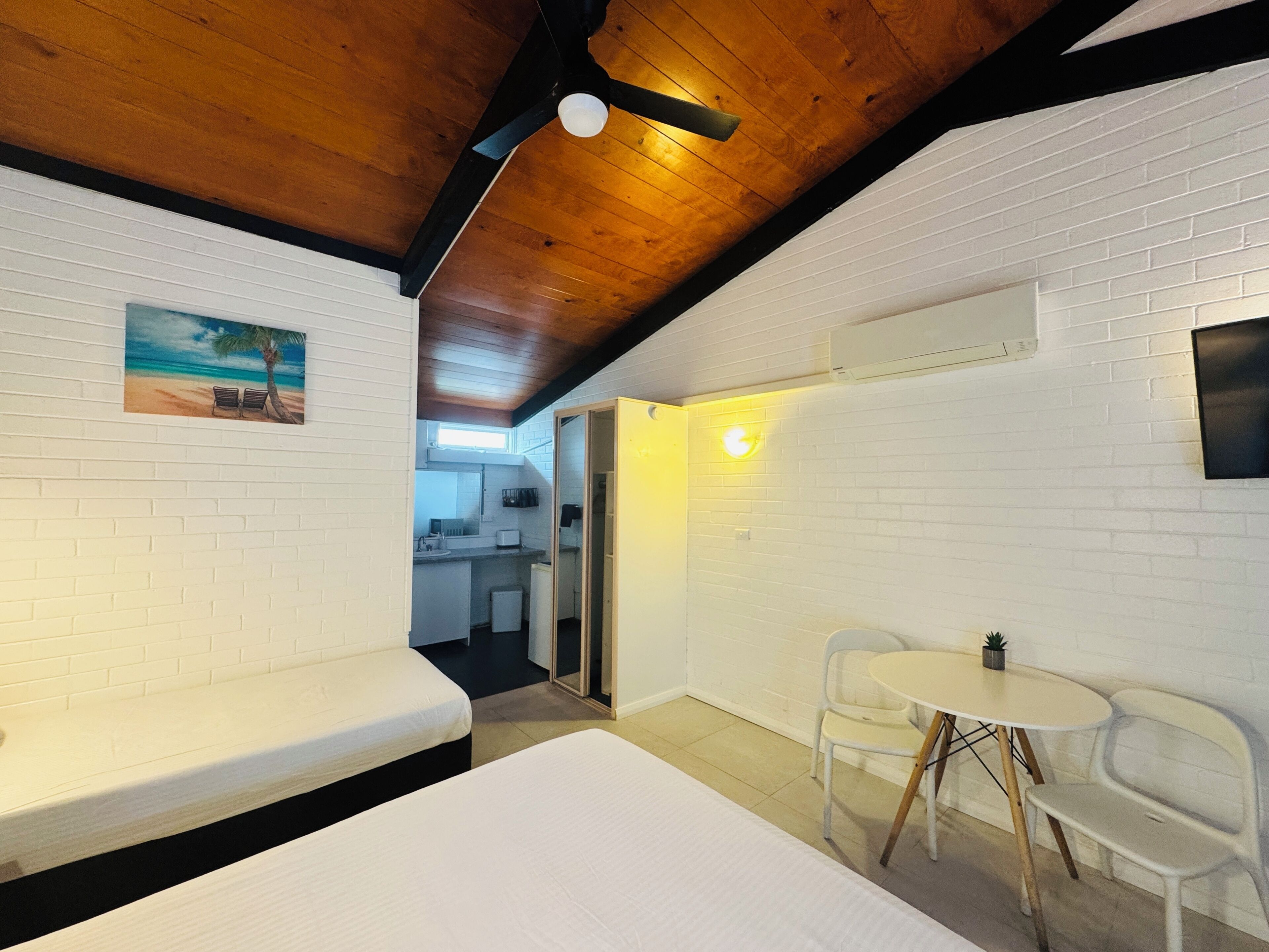 Een kamer met een houten plafond, witte bakstenen muren, een bed, een bank, een kleine tafel met stoelen en een schilderij van een strand.