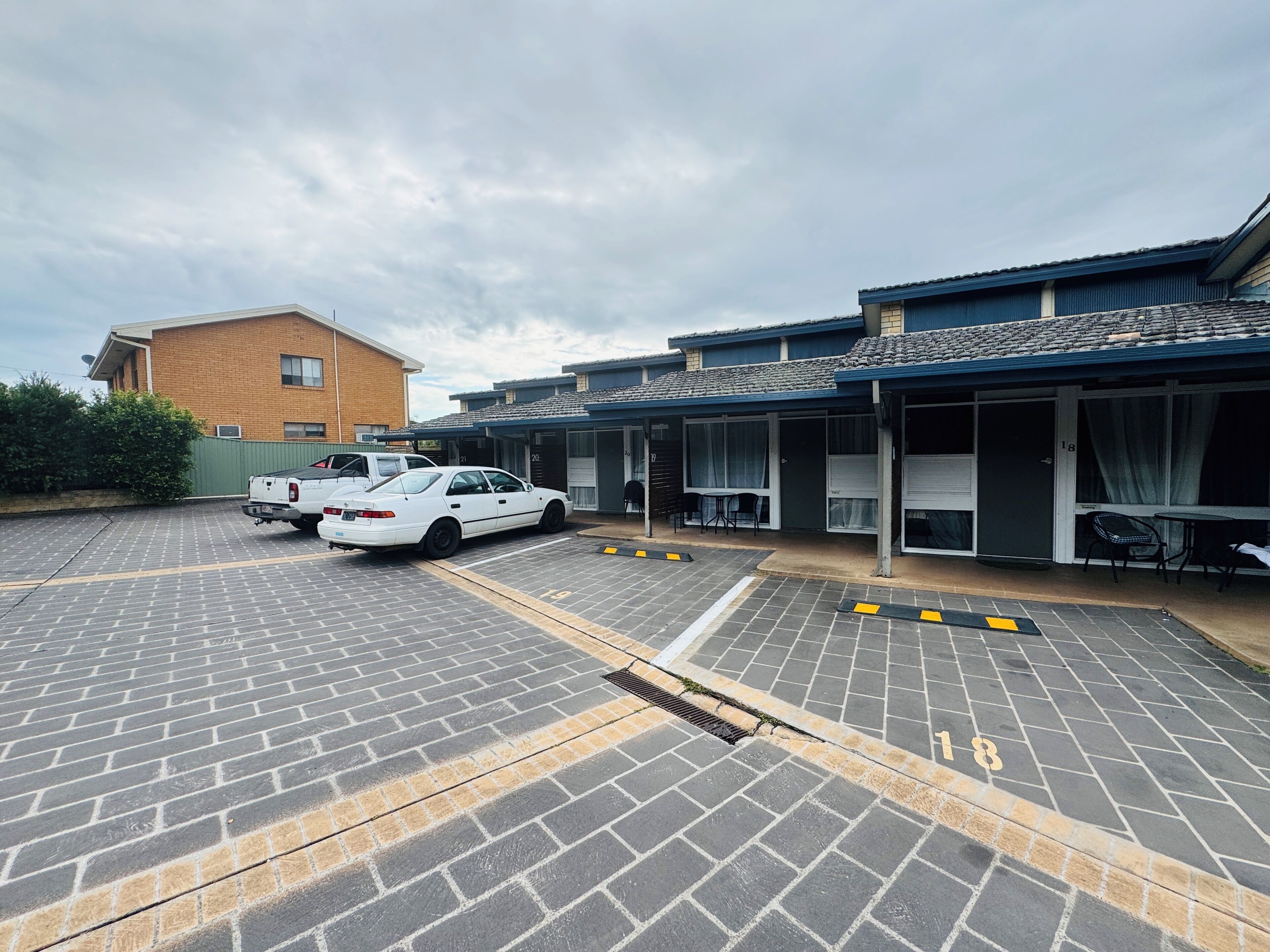 A parking lot with two white cars, a brick building, and a row of rooms with sliding doors.