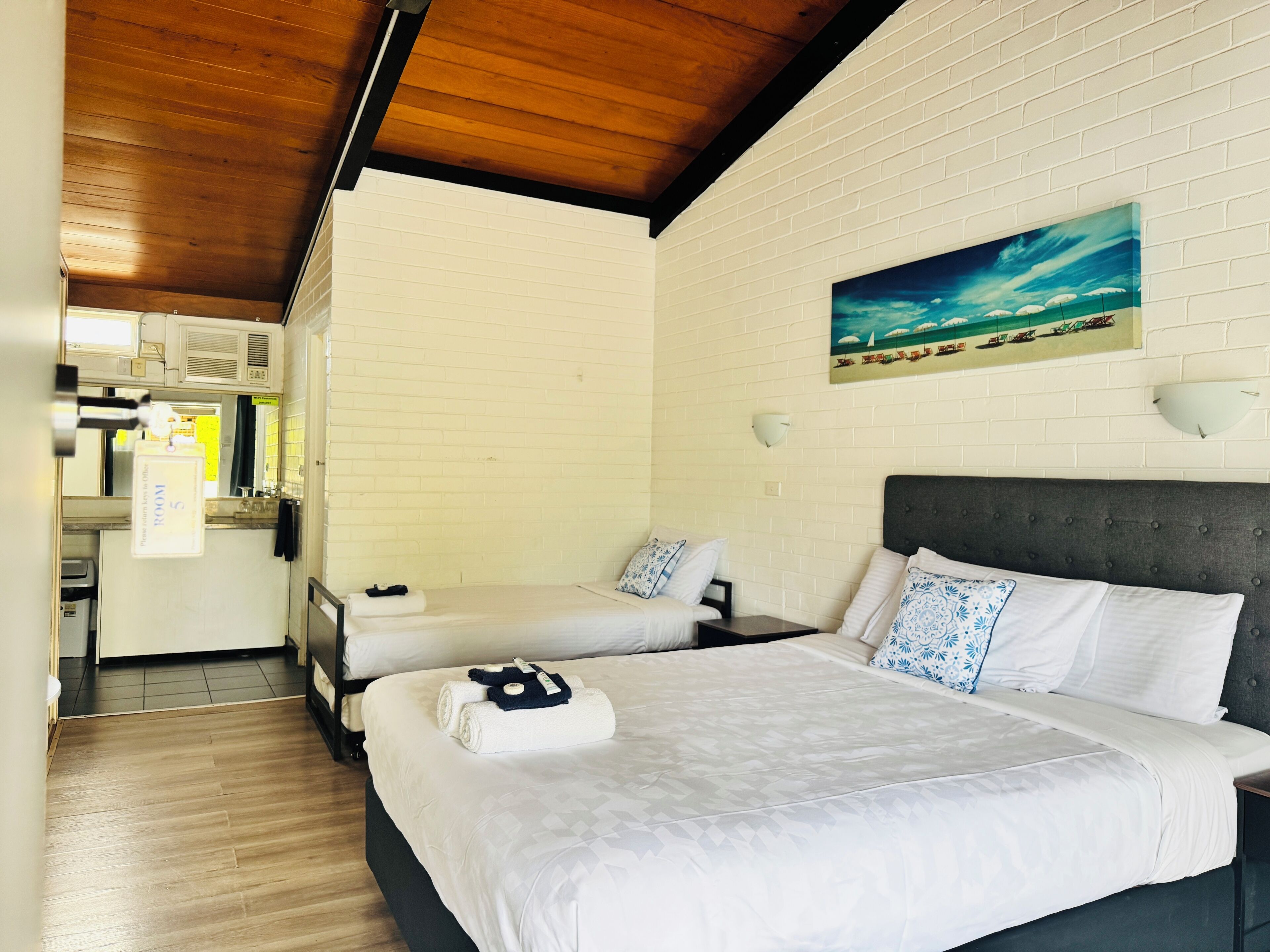 Una habitación de hotel con una cama grande, una cama más pequeña, un cuadro con una playa y una zona de kitchenette.