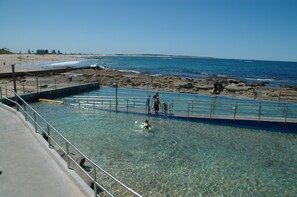 A swimming pool with clear water, a beach in the background, and a rocky shoreline.
