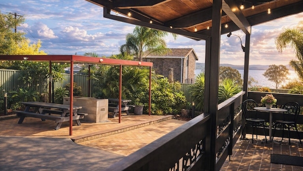 An outdoor patio with a covered seating area, a stone fireplace, and a view of the sea. - Jetty Motel The Entrance (Long Jetty)
