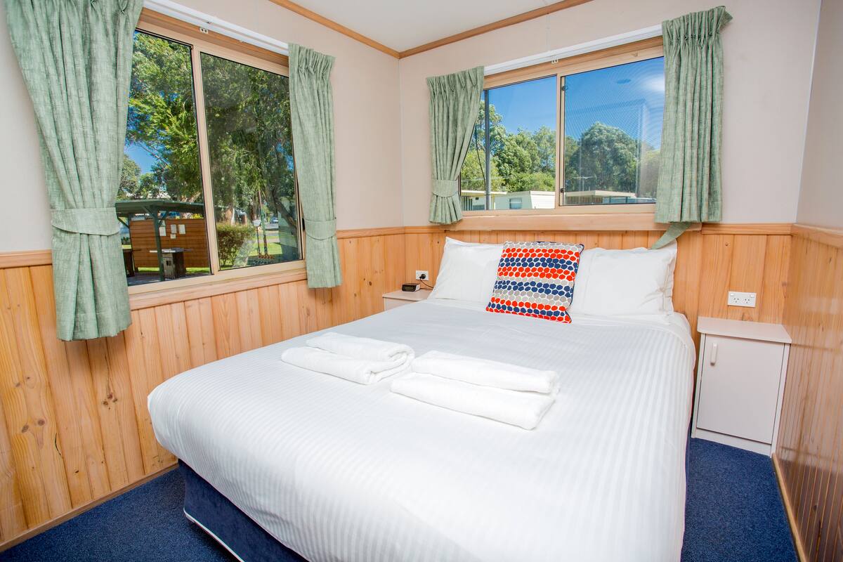 A neatly made bed with white linens and two folded towels, a wooden headboard, a small bedside table, and a window with curtains.