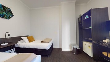 Habitación de hotel con cama, escritorio con lámpara, armario, cubo de basura y refrigerador pequeño.