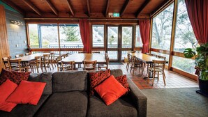 A cozy indoor seating area with a sofa, red cushions, wooden tables, and chairs. Large windows provide a view of the outdoors. - YHA Thredbo (Thredbo)