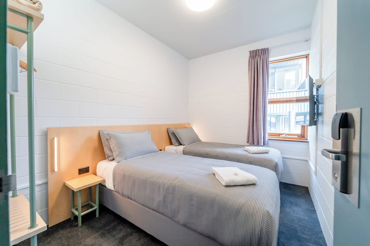 A small, clean room with two beds, a small table, a window with curtains, and a wall-mounted TV.