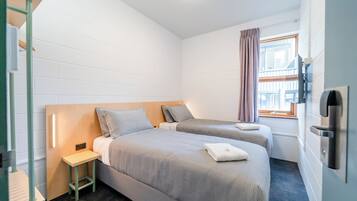 Quarto pequeno e limpo, com duas camas, uma mesinha, janela com cortinas e televisão na parede.