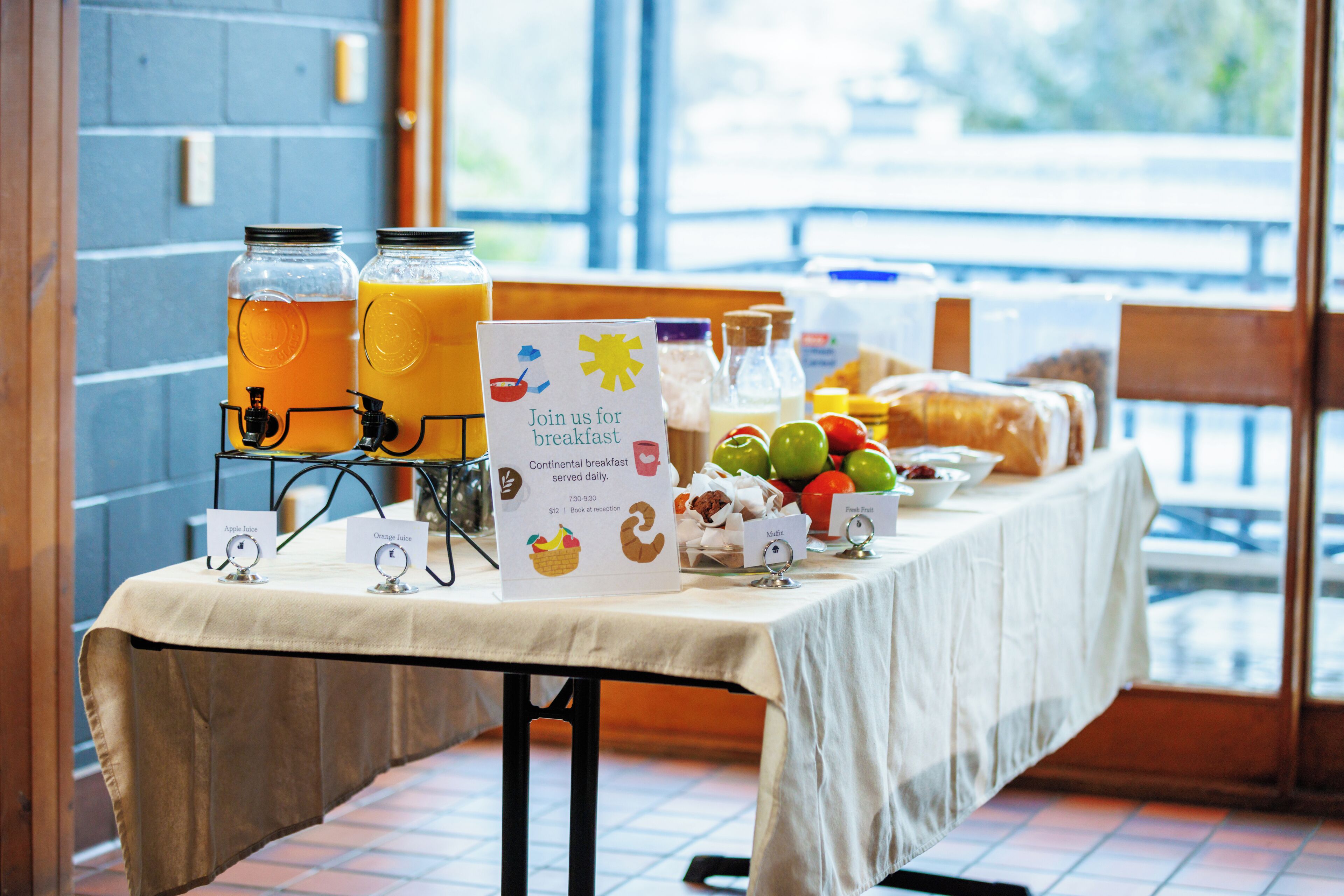 A breakfast table with juice dispensers, a sign inviting guests to join for breakfast, and a variety of fruits and pastries.