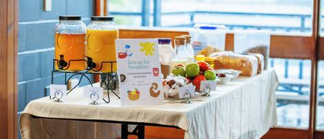 Una mesa de desayuno con dispensadores de jugo, un cartel invitando a los huéspedes a desayunar, y una variedad de frutas y pasteles.