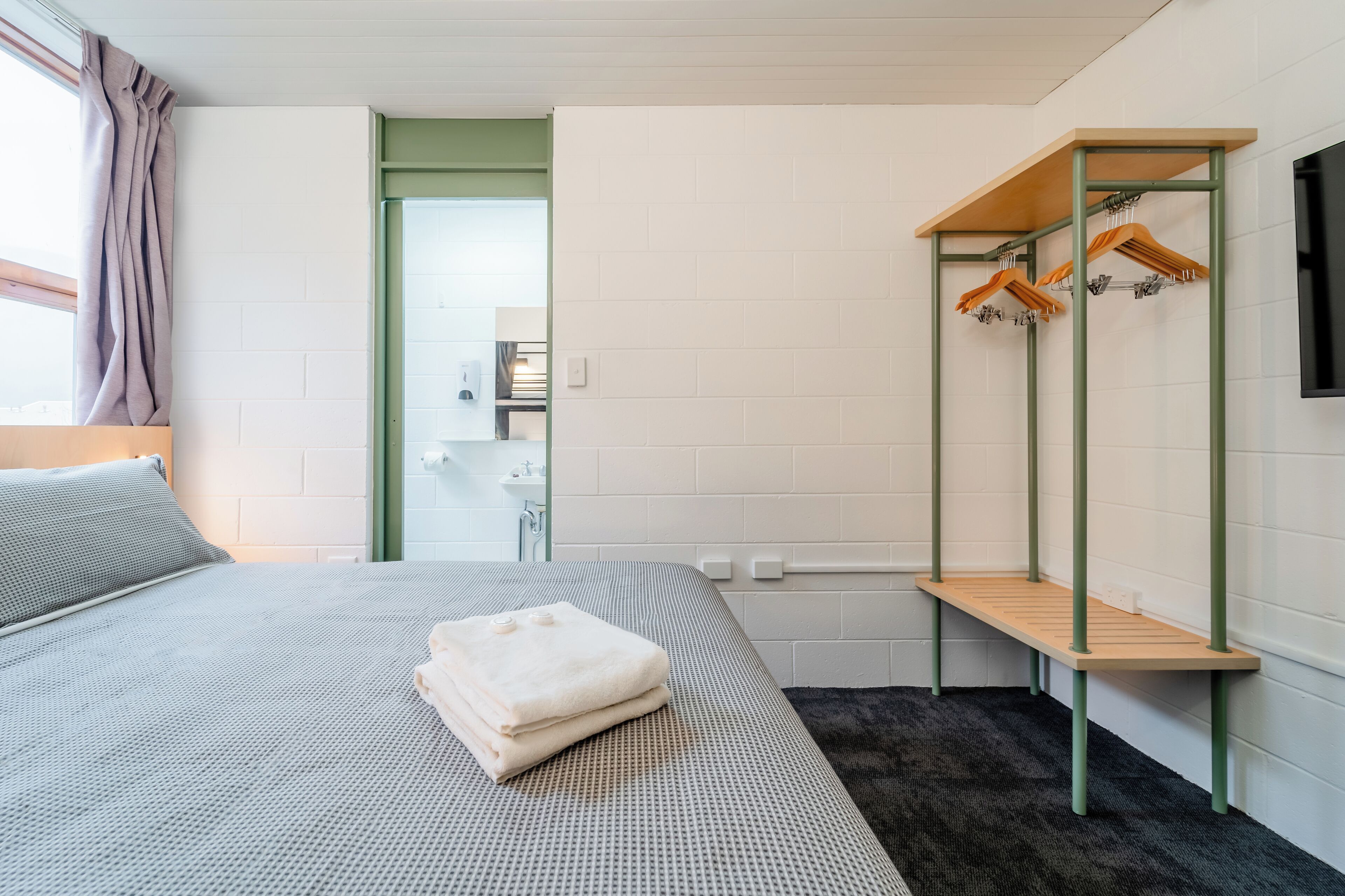 Habitación de hotel moderna con cama, un banco de madera con ropa colgando y vista al baño.