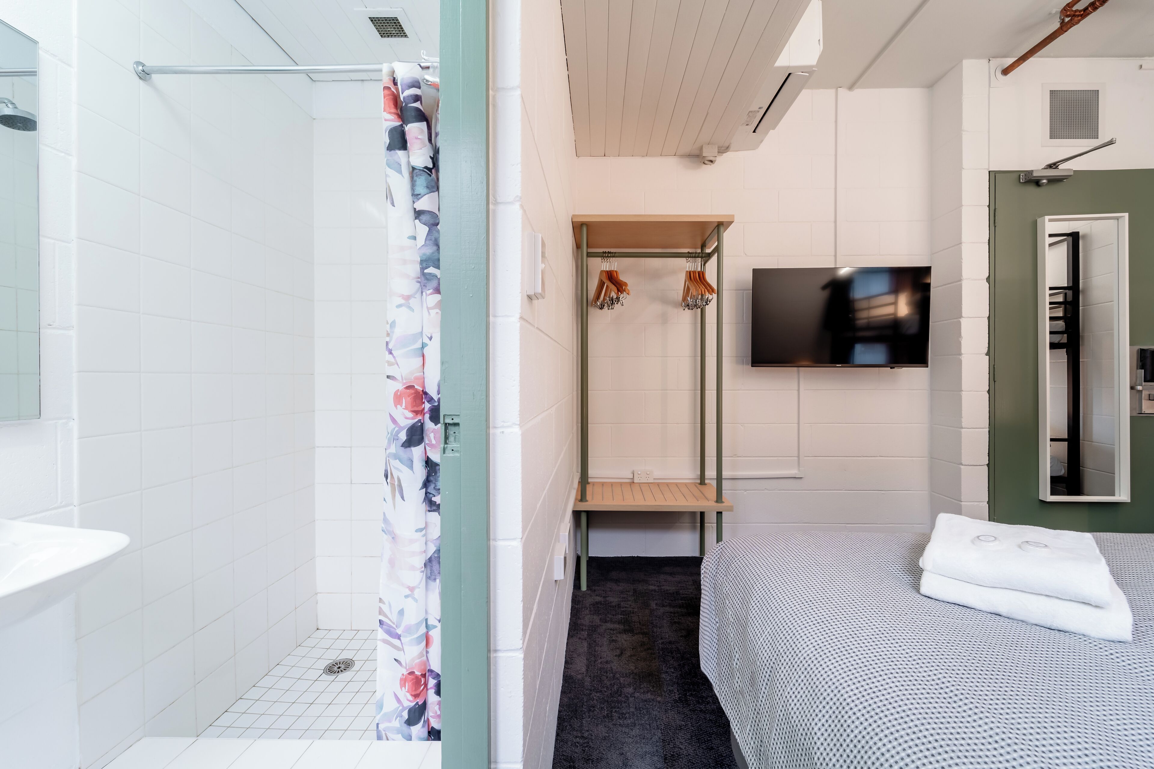 Un baño moderno con azulejos blancos, una ducha con cortina estampada de flores, un banco de madera, un televisor montado en la pared y una cama con colcha estampada.