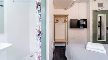 Un baño moderno con azulejos blancos, una ducha con cortina estampada de flores, un banco de madera, un televisor montado en la pared y una cama con colcha estampada.
