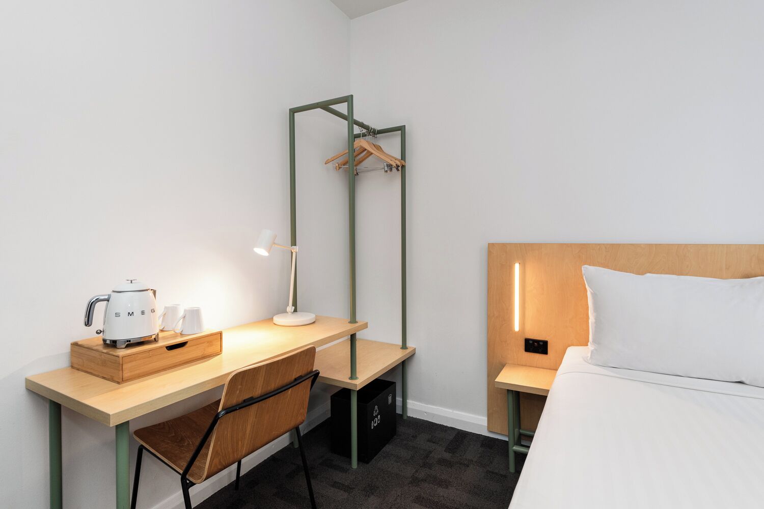 A hotel room with a wooden desk, a chair, a bed with a wooden headboard, a hanging coat rack, and a bedside table with a lamp.