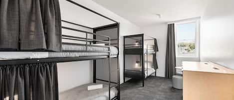 A dormitory-style room with bunk beds, a desk, and a window with curtains.