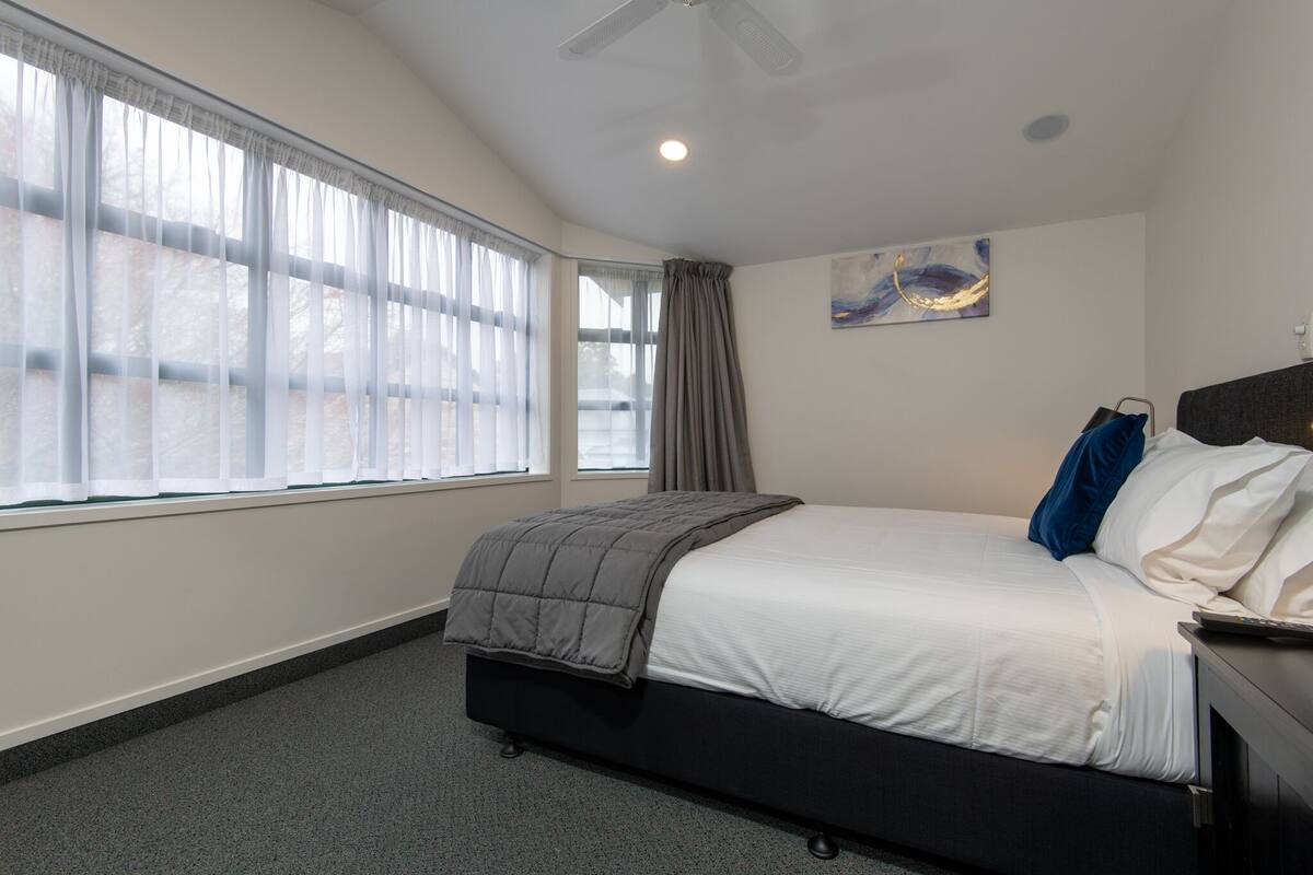 A bedroom with a bed, a window with curtains, a ceiling fan, and a painting on the wall.