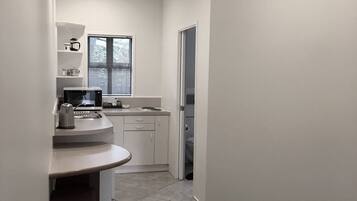 A compact kitchen with a countertop, a kettle, and a window with blinds.