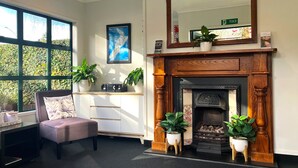 A room with a fireplace, a chair, a desk, and a large window with a view of greenery. - Cambridge Mews (Cambridge)