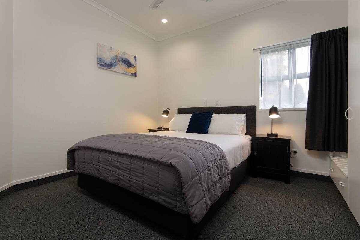 A neatly arranged bedroom with a bed, bedside tables, a window with curtains, and a painting on the wall.