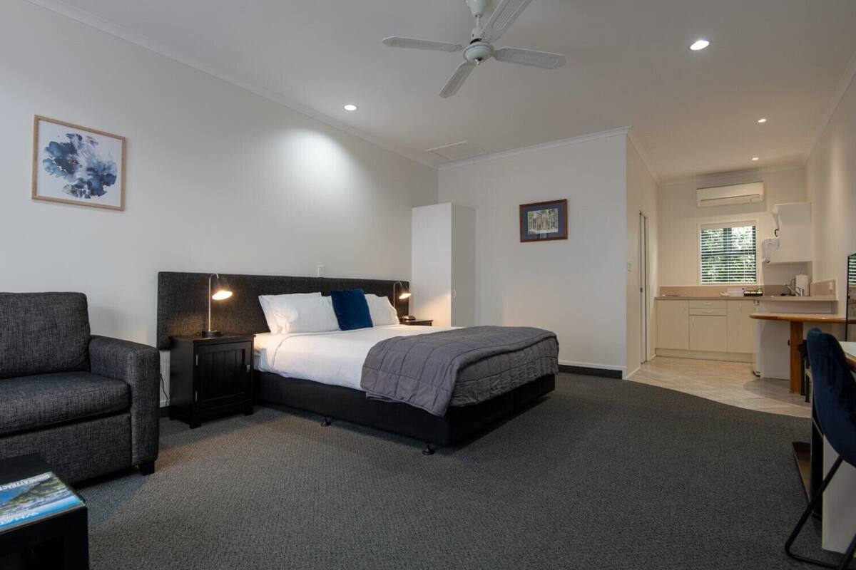 A hotel room with a bed, a desk, a chair, a nightstand, a ceiling fan, and a kitchenette.