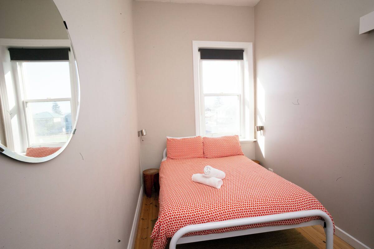 A small, single-bed room with a round mirror, two windows with blinds, and a neatly made bed with a red and white bedspread.