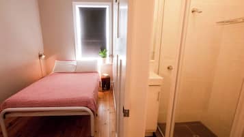 A bedroom with a bed, a window, a bedside table with a plant, and a shower area.