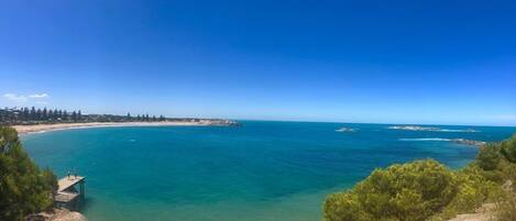 A coastal landscape with clear blue waters, a sandy beach, and a rocky outcrop with a viewing platform.