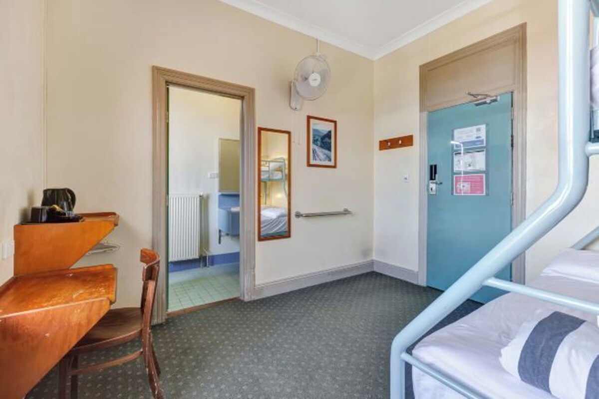 A hotel room with a bed, a desk, a chair, a door, and a mirror.