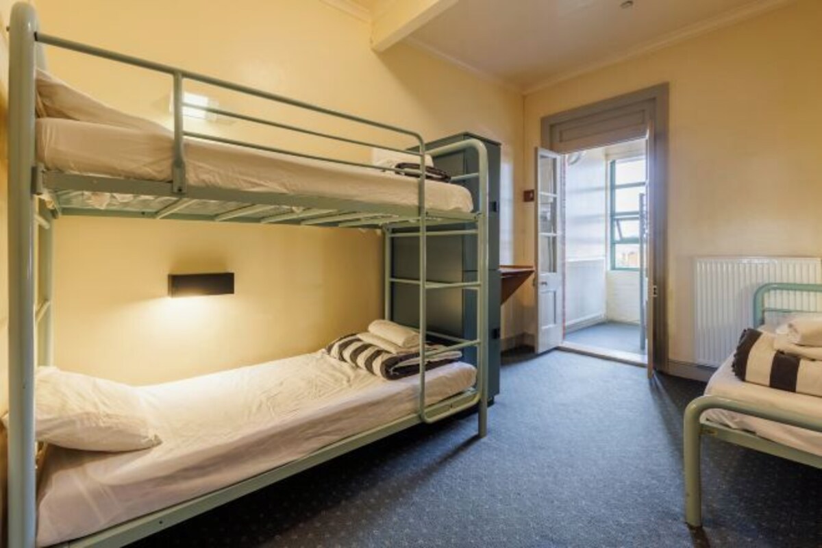 A dormitory room with bunk beds, a small window, and a door leading to a balcony.