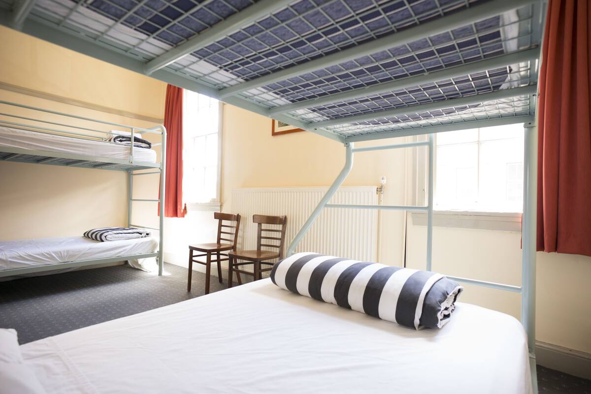 A dormitory room with bunk beds, a desk, chairs, and a window with curtains.
