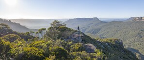 Una persona está de pie sobre un promontorio rocoso, con una vista panorámica de un paisaje montañoso.