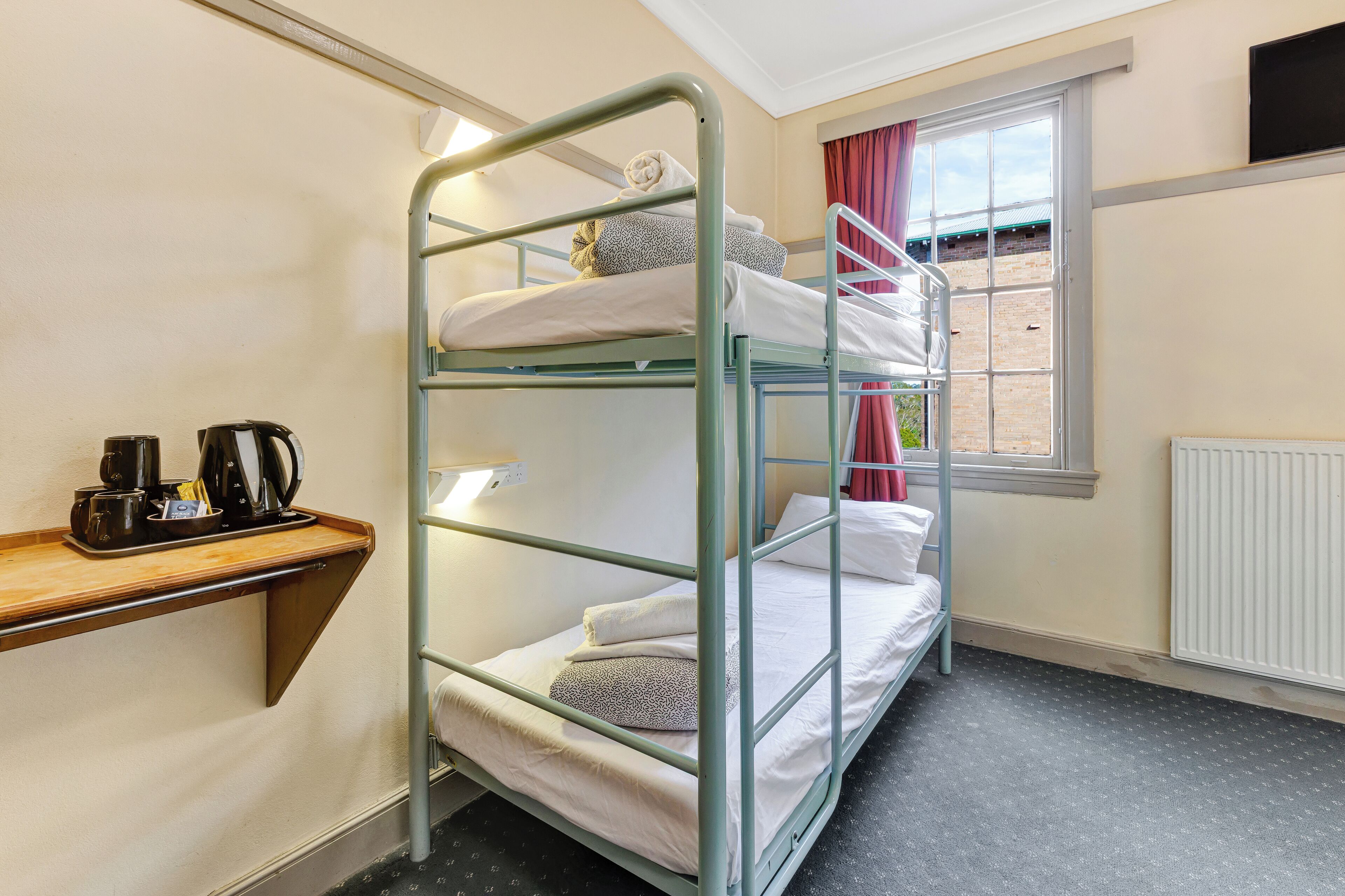Een slaapkamer met een stapelbed, een houten plank met een koffiezetapparaat en bekertjes, een raam met gordijnen en een radiator.