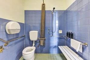 A bathroom with blue tiles, a white toilet, a shower with a grab bar, and a bench.