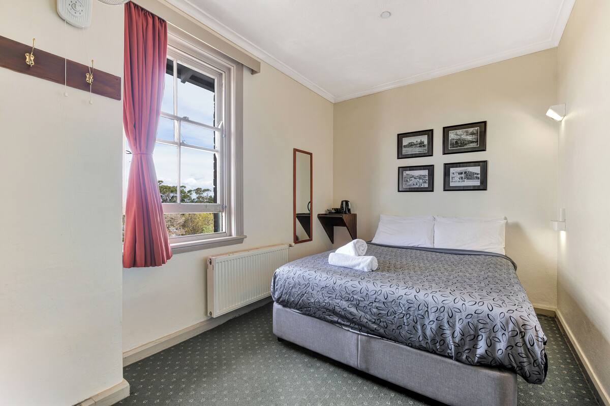 A neatly arranged bedroom with a bed, a window with curtains, a mirror, and framed pictures on the wall.