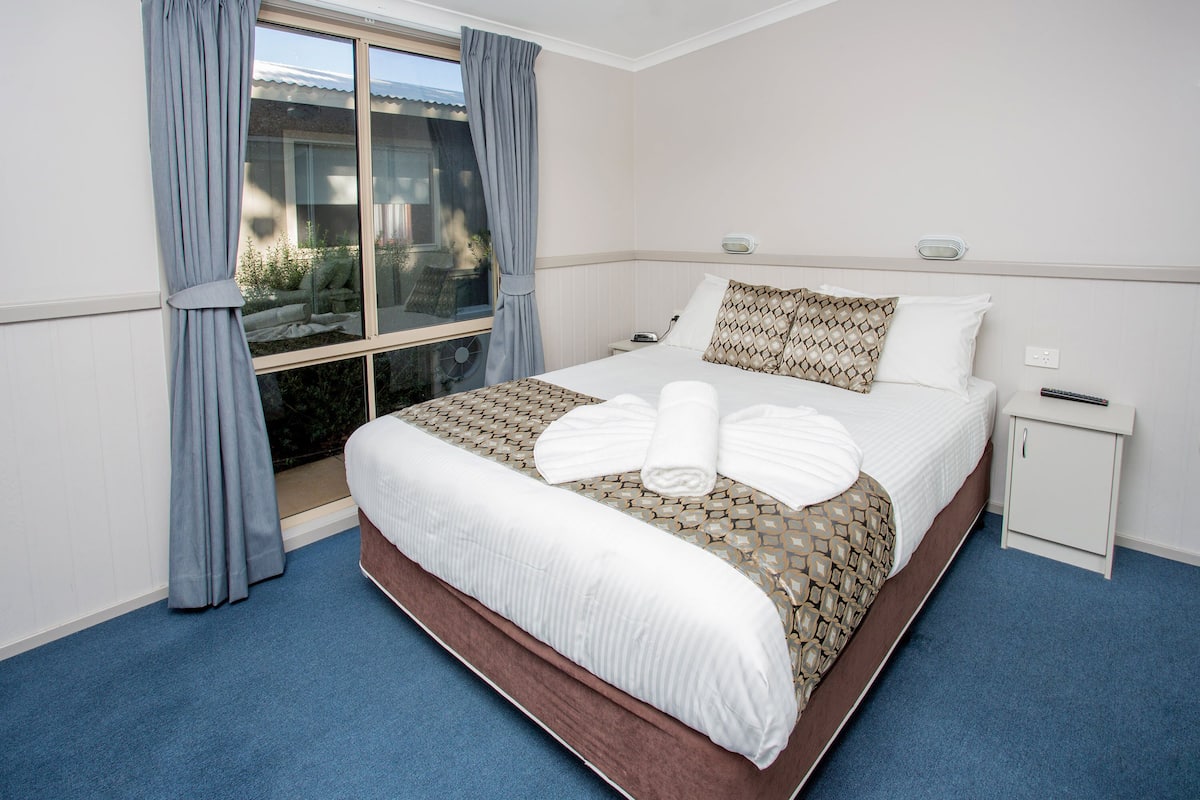 A neatly made bed with white linens and a patterned bedspread, a bedside table with a lamp, and a window with curtains.