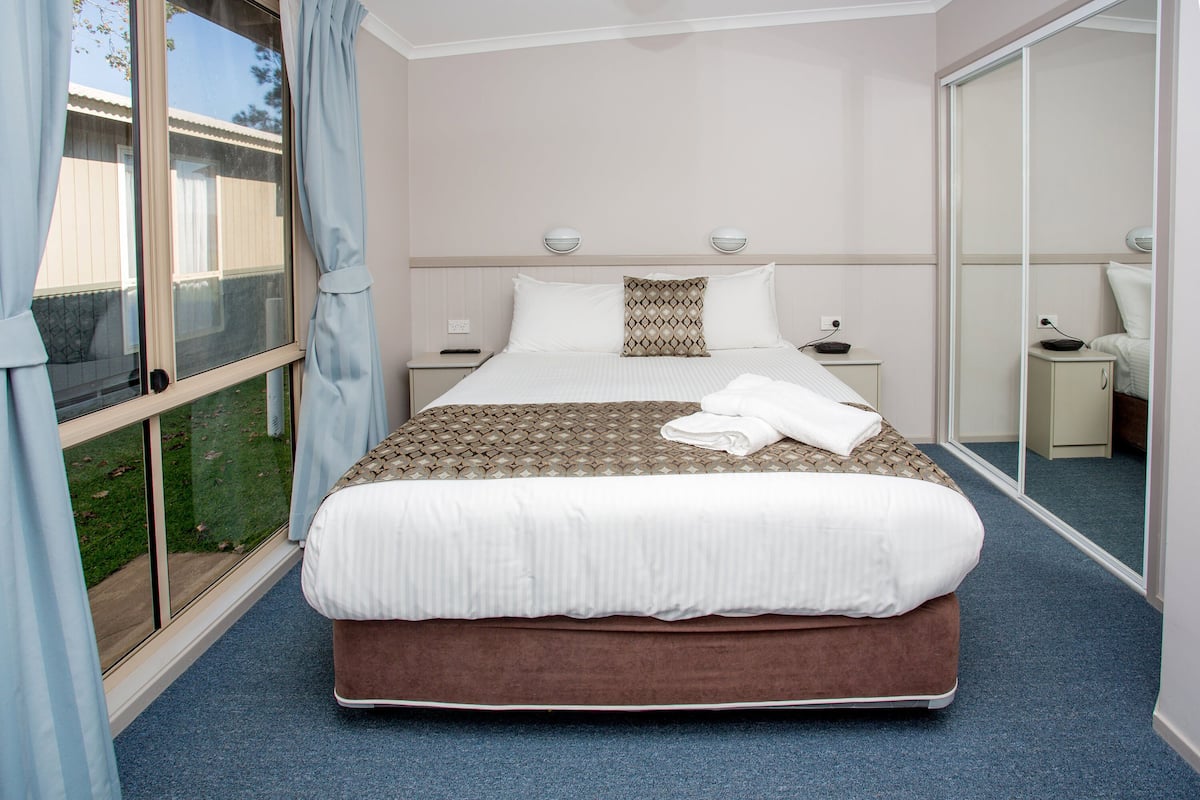 A hotel room with a large bed, a window with curtains, a bedside table with a lamp, and a small bedside table with a bowl.
