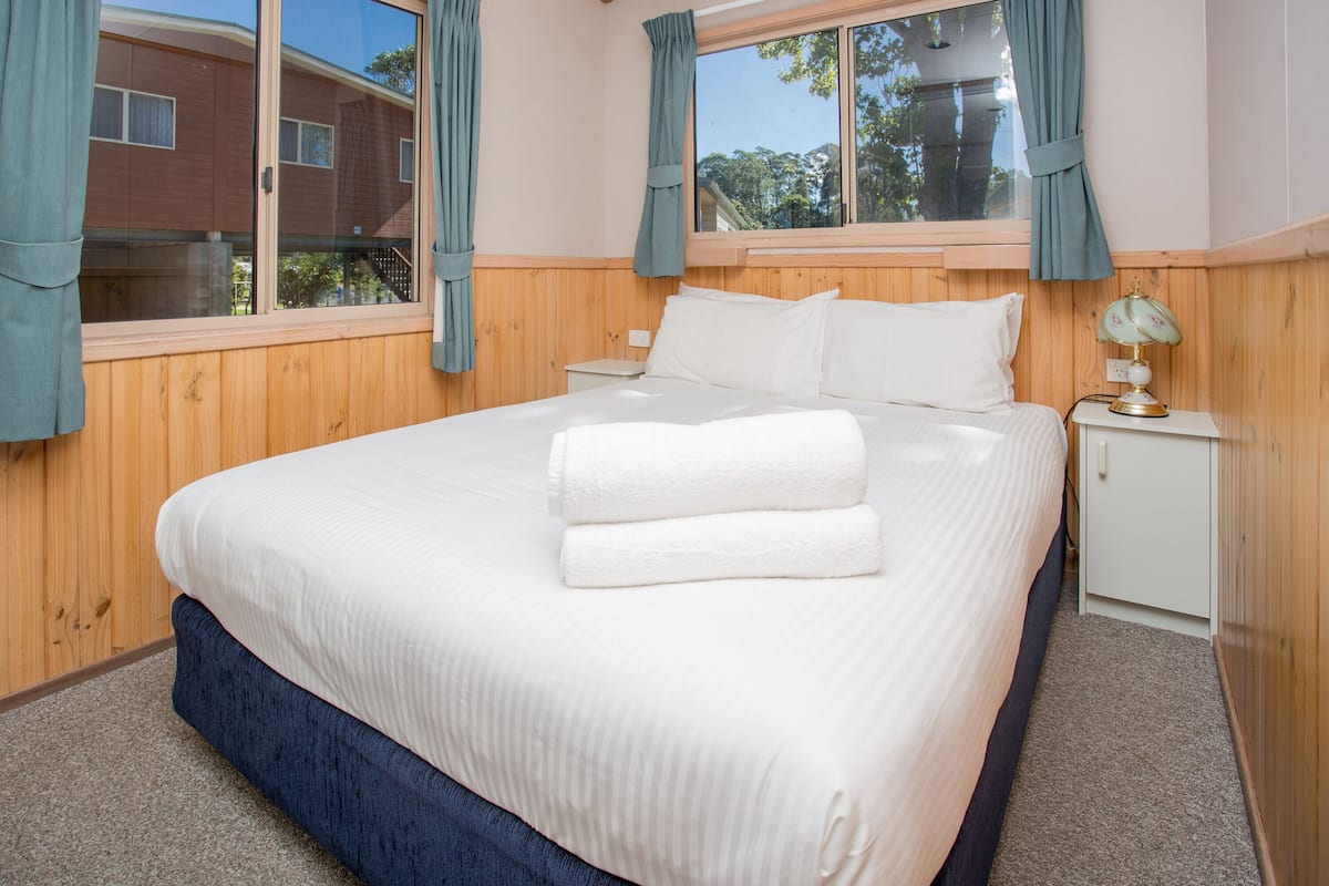 A neatly made bed with white linens and two folded towels, a bedside table with a lamp, and a window with curtains.