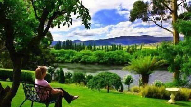 A person sitting on a bench in a lush garden with a river and mountains in the background. - The Shingles Riverside Cottages (New Norfolk)