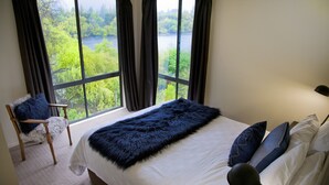 A bedroom with a large window, a bed with white linens, a blue furry throw, and a chair with a blue cushion. - The Shingles Riverside Cottages (New Norfolk)
