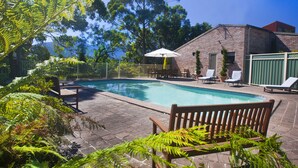 A swimming pool with lounge chairs and an umbrella, surrounded by greenery and a brick building. - The Shingles Riverside Cottages (New Norfolk)