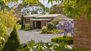 A two-story brick house with a covered porch, surrounded by well-maintained gardens and trees. - The Shingles Riverside Cottages (New Norfolk)