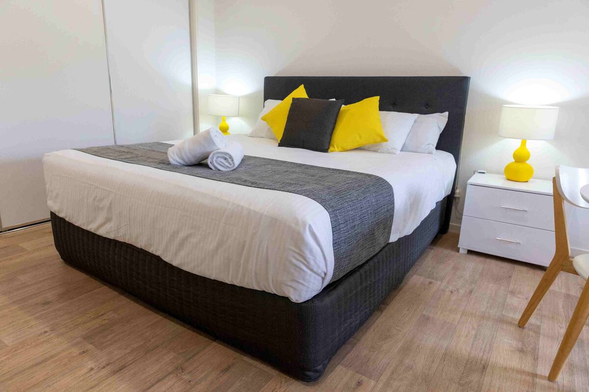 A neatly made bed with a grey and white duvet, two pillows, and a rolled towel on the side. There are two bedside tables with lamps, one of which has a yellow lamp base. The room has a wooden floor and white walls.