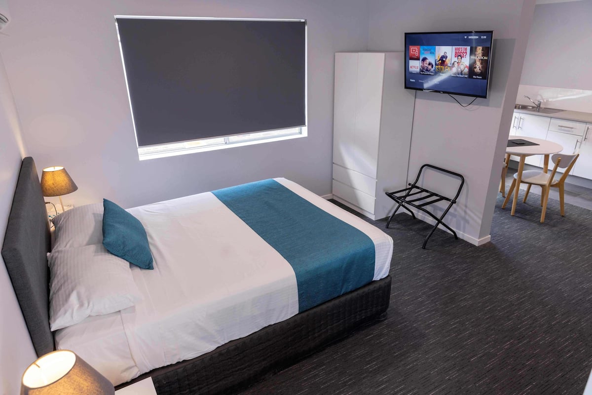 A hotel room with a bed, a TV mounted on the wall, a desk with a chair, and a window with a blind.