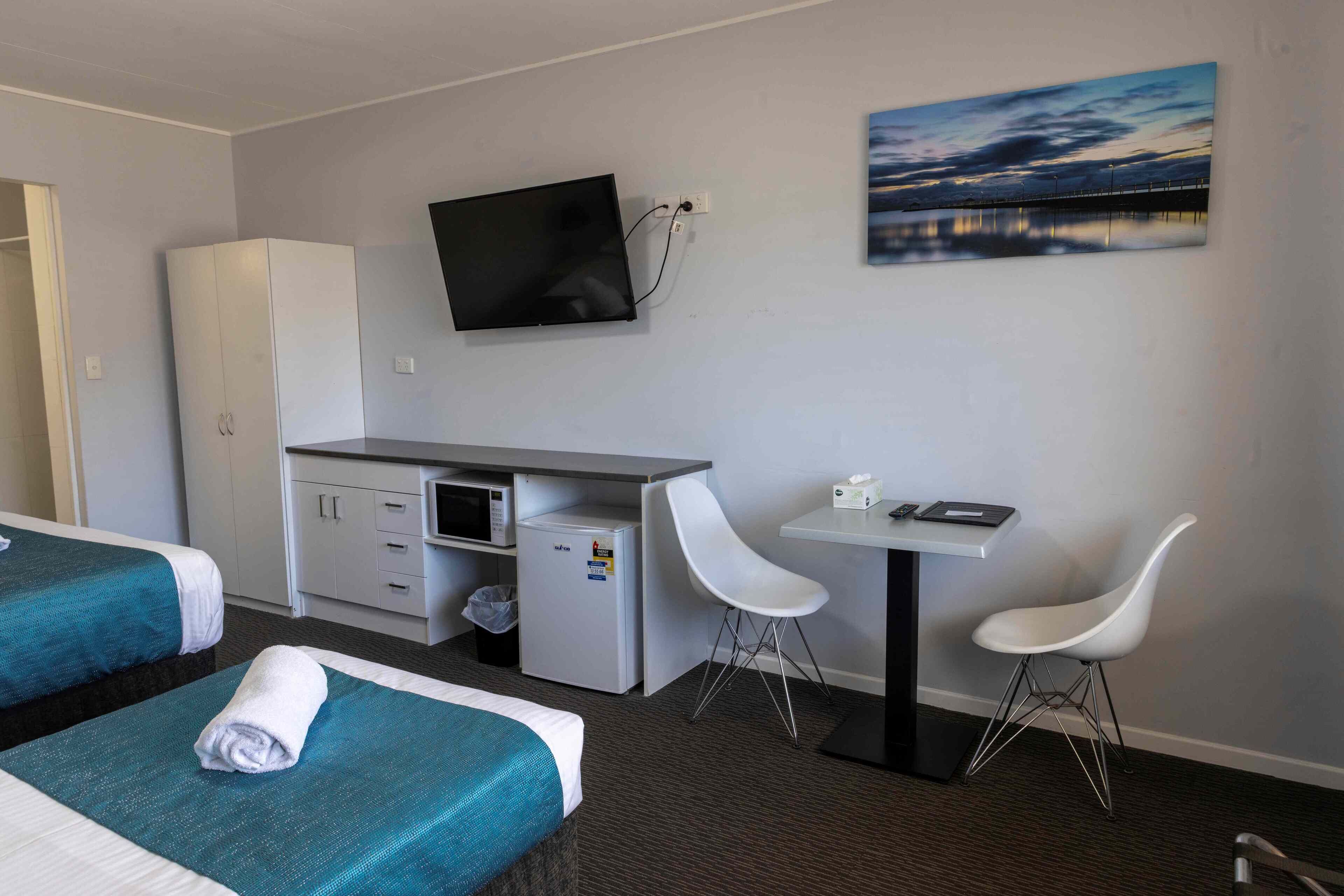 A hotel room with two beds, a desk, a TV, a refrigerator, and two white chairs.
