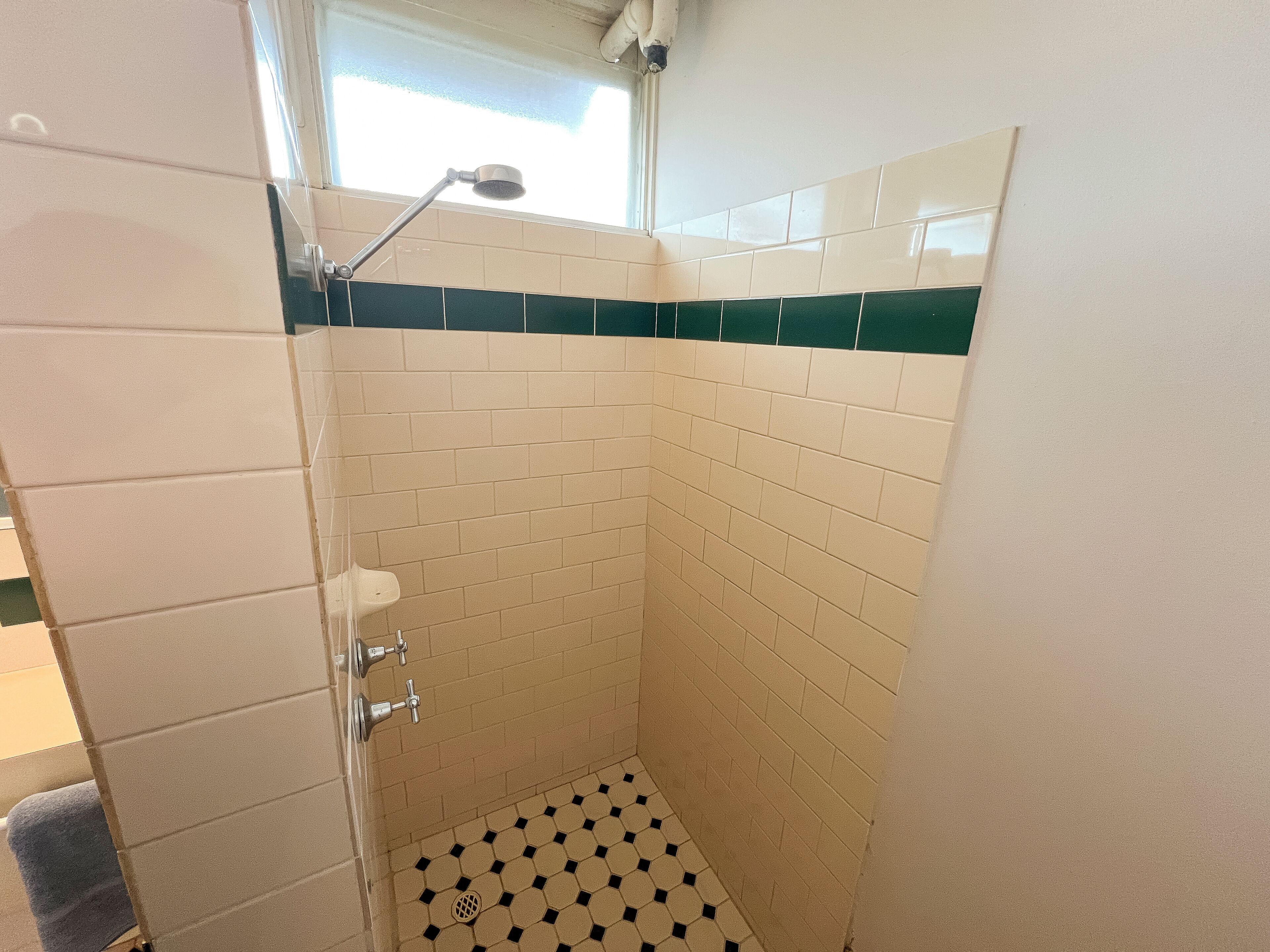 Une petite douche carrelée, dotée d’un pommeau de douche à main, d’un drain visible et d’une fenêtre située au-dessus.