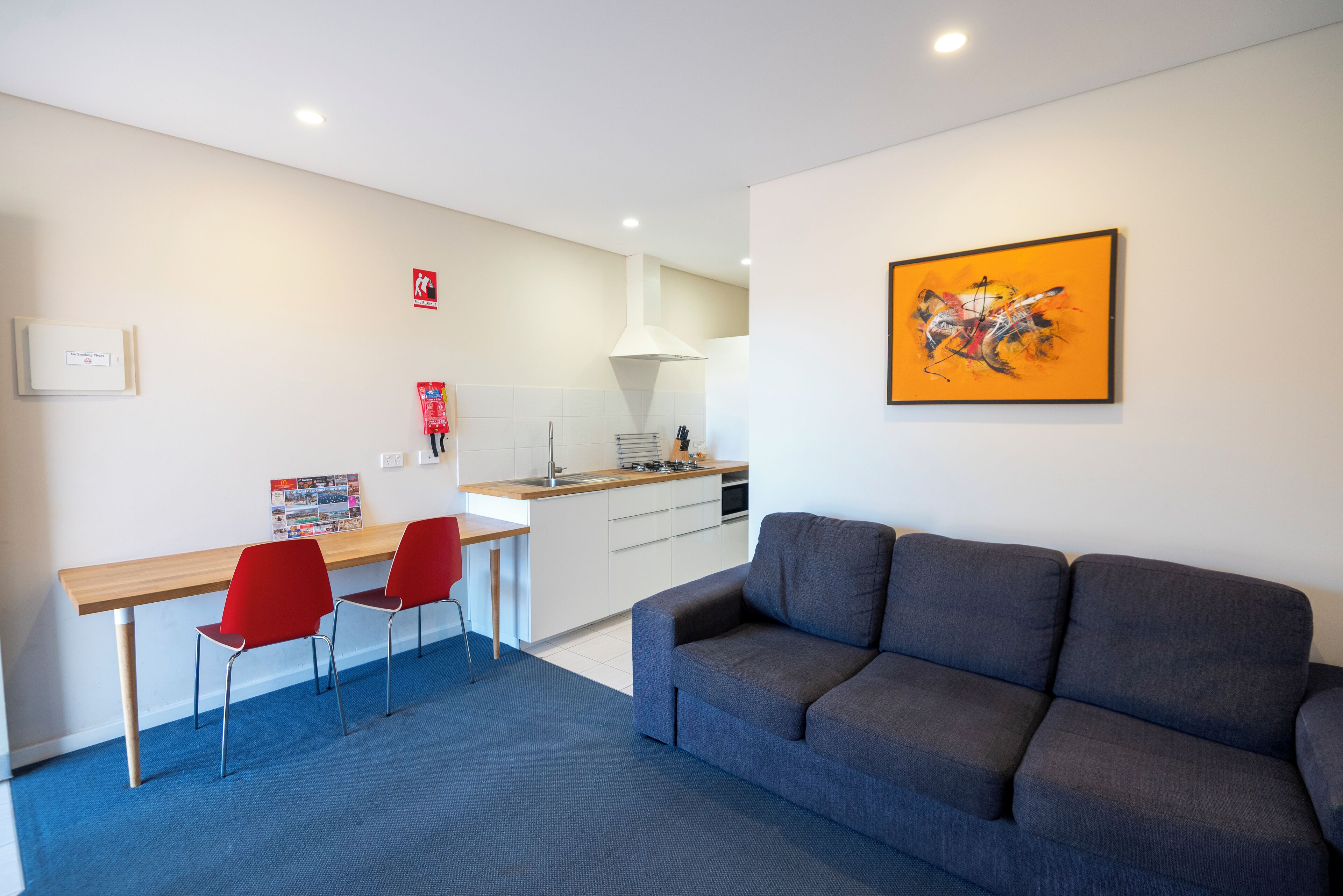 Un salon moderne avec un canapé gris, un bureau en bois avec deux chaises rouges, et un coin cuisine à l’arrière-plan.