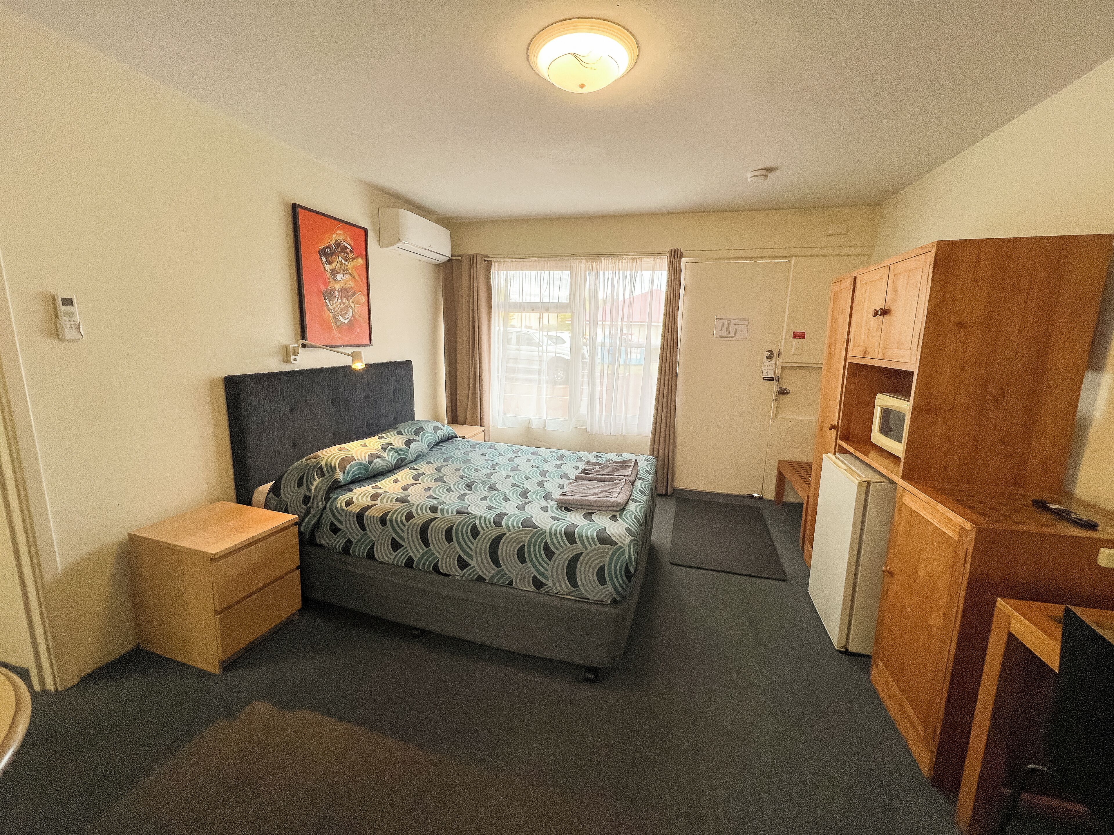 A hotel room with a bed, bedside table, wardrobe, and a small desk with a chair.