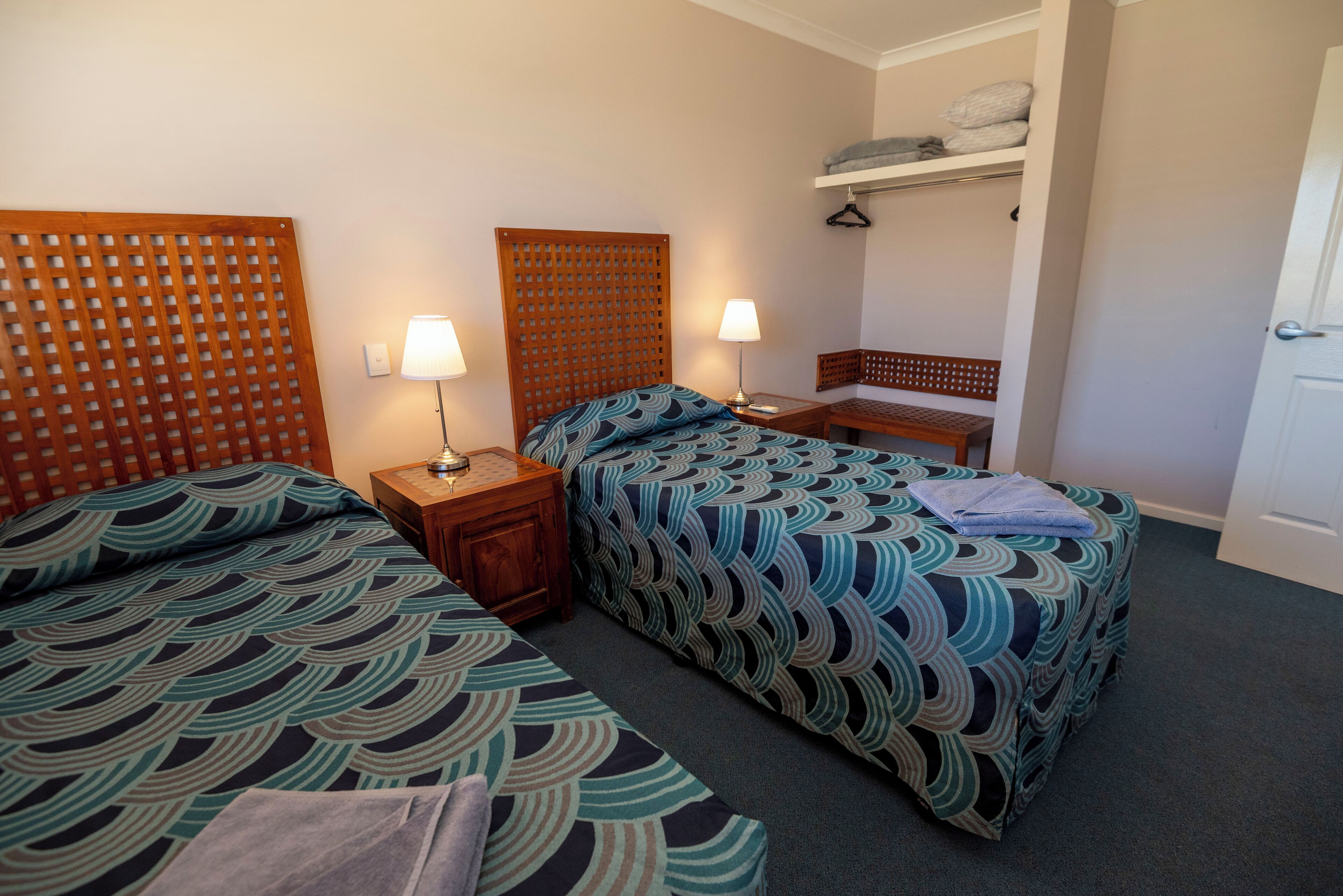 A hotel room with two beds, each with a patterned bedspread, a wooden headboard, a bedside table with a lamp, and a small bench.