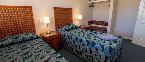 Habitación de hotel con dos camas, cada una con colcha estampada, cabecero de madera, mesita de noche con lámpara y un pequeño banco.