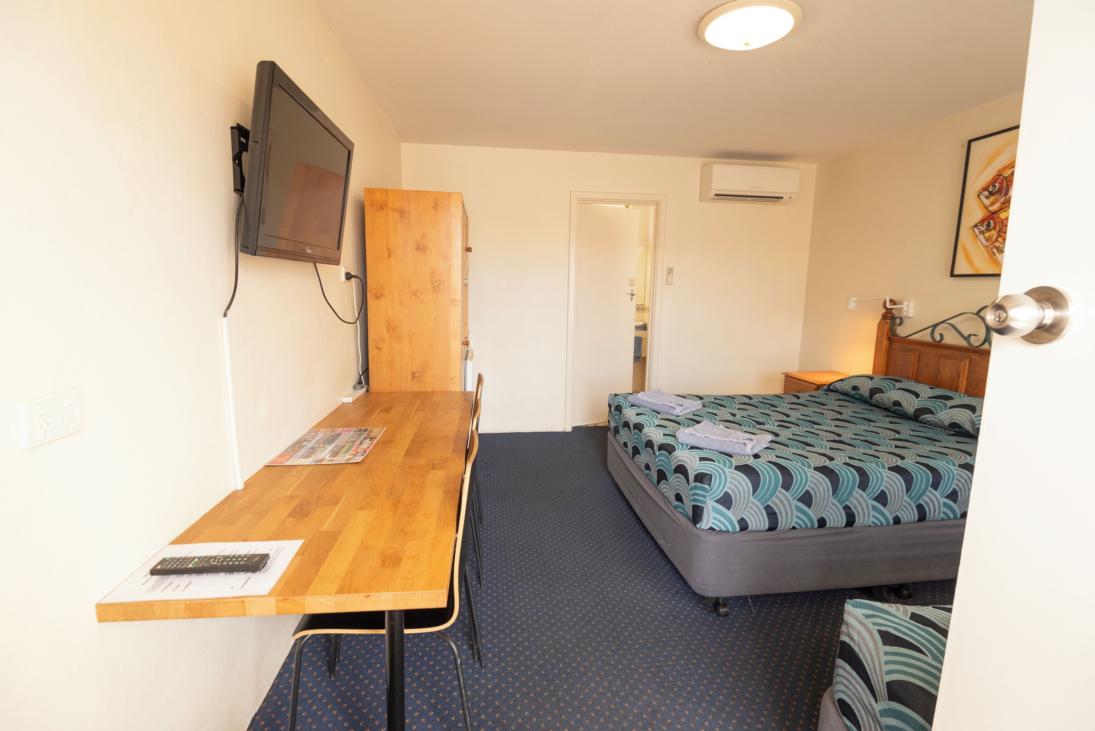 A small, compact room with a wooden desk, a bed with patterned bedding, a television mounted on the wall, and a door leading to another room.