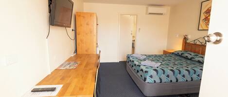 A small, compact room with a wooden desk, a bed with patterned bedding, a television mounted on the wall, and a door leading to another room.